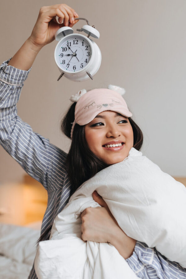 Cute girl holding alarm clock above her head and with smile hugging white pillow